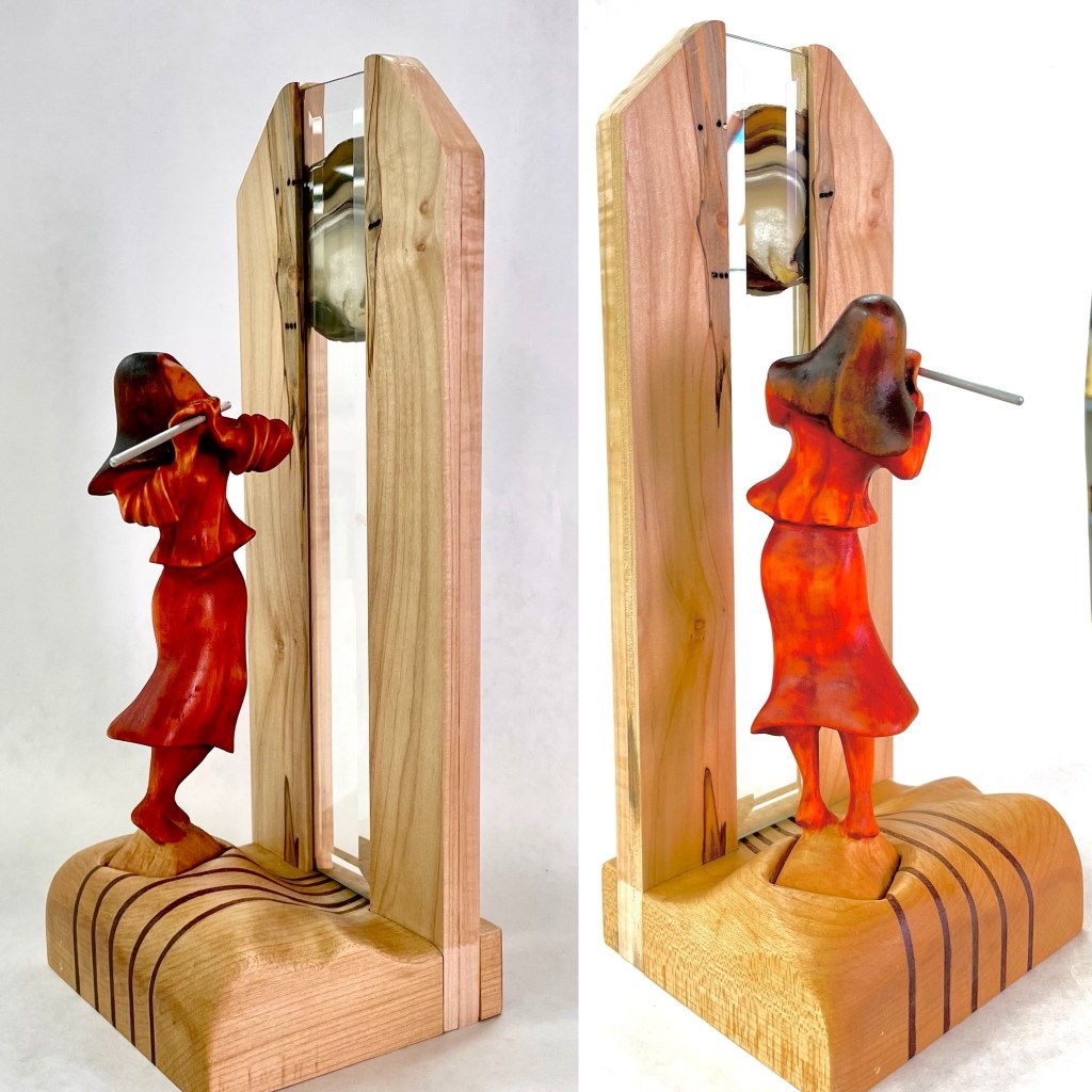 A wooden sculpture featuring a young woman playing a flute, positioned between two upright, arch-like wooden panels with a glass bevel, symbolizing the transition between dreams and reality.